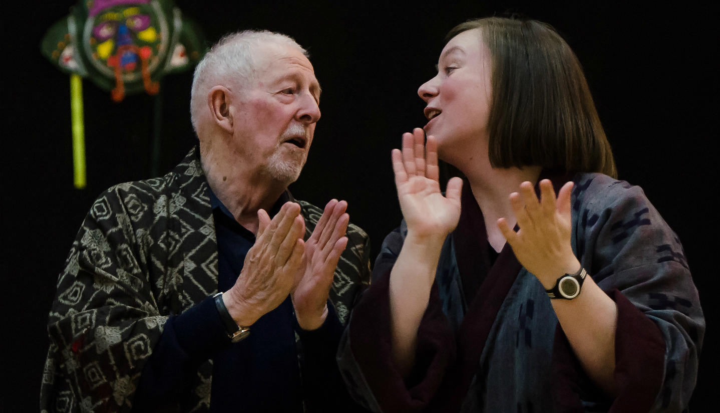 An older man and a younger woman sing facing each other in a Memory Spinners performance of The Mikado. They are clapping and wearing dark robes.